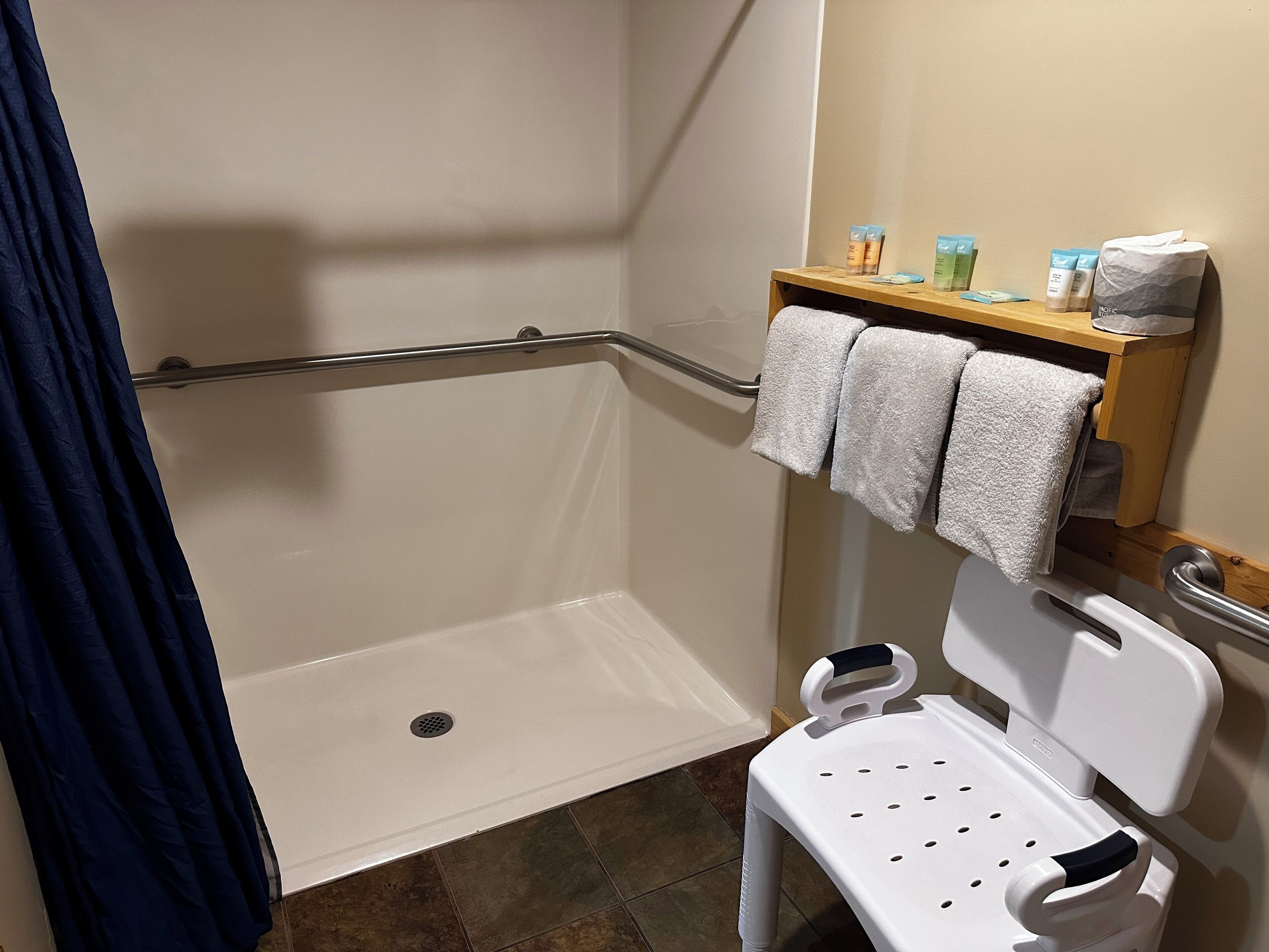 Un espace douche équipé d’une barre d’appui, d’une chaise blanche et d’une étagère contenant des articles de toilette.