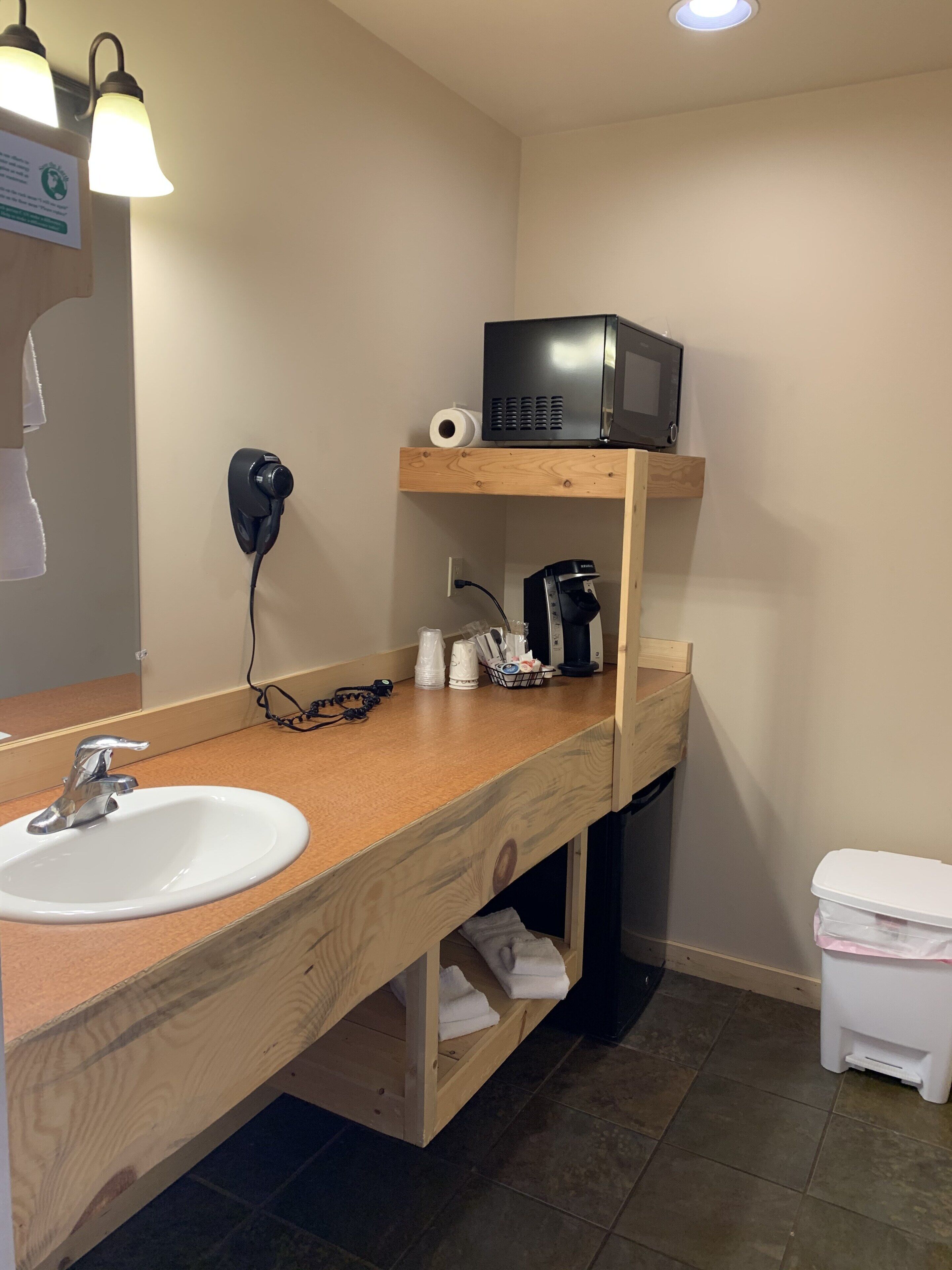 A bathroom with a wooden vanity, a sink, a mirror, a microwave, a coffee maker, and a trash bin.