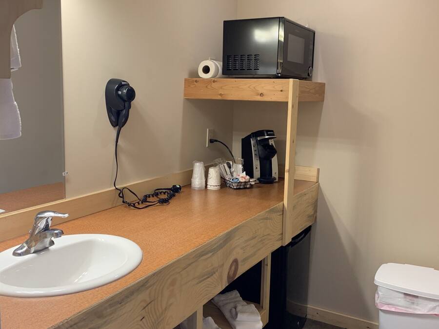 A bathroom with a wooden vanity, a sink, a mirror, a microwave, a coffee maker, and a trash bin.