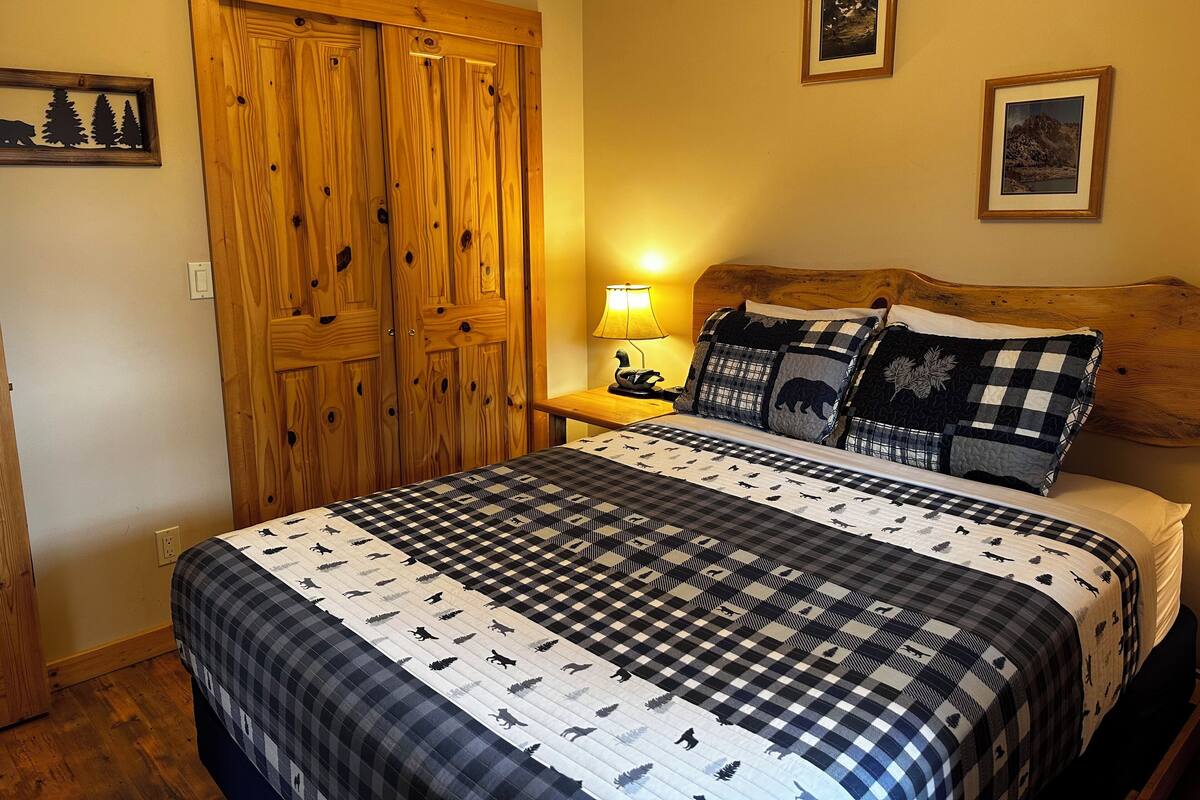 A bedroom with a bed, wooden wardrobe, two framed pictures on the wall, and a nightstand with a lamp.
