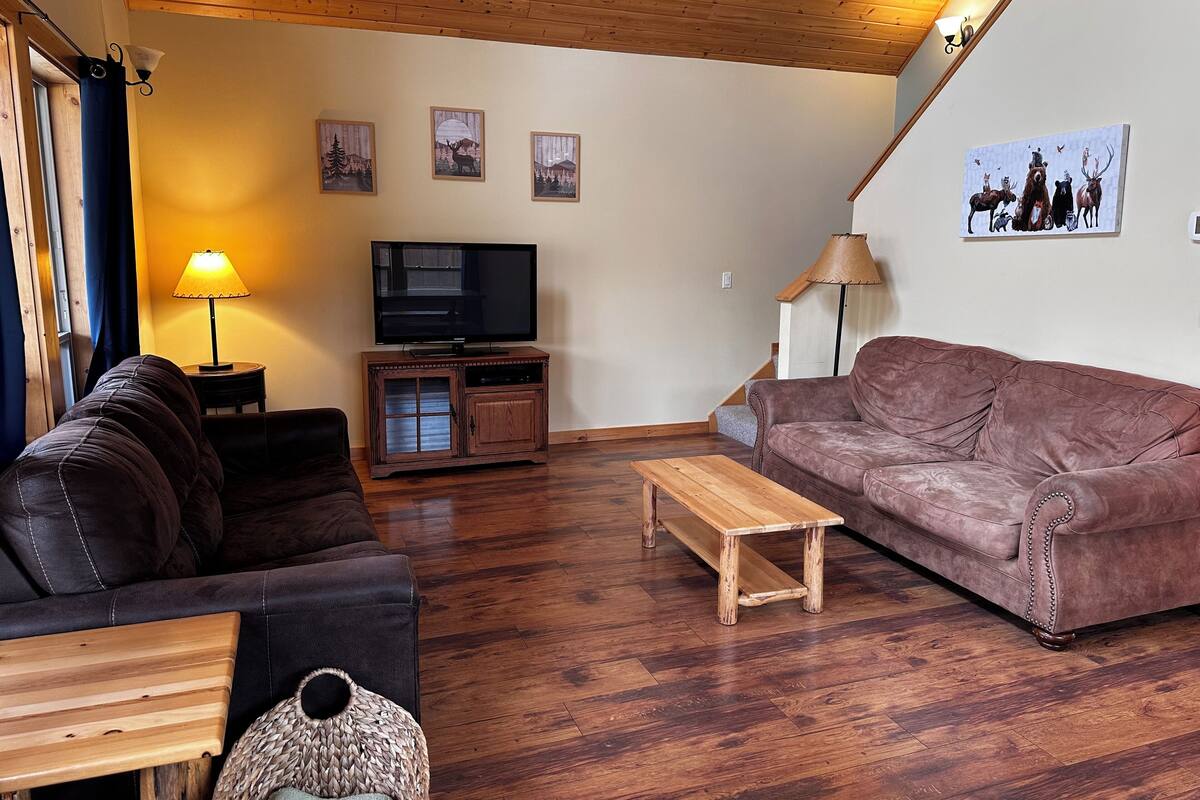 A living room with a brown leather couch, a wooden coffee table, a TV on a wooden stand, and a staircase leading upstairs.