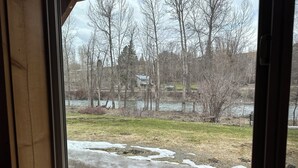 A view through a window of a river, trees, and a grassy area with some snow patches.