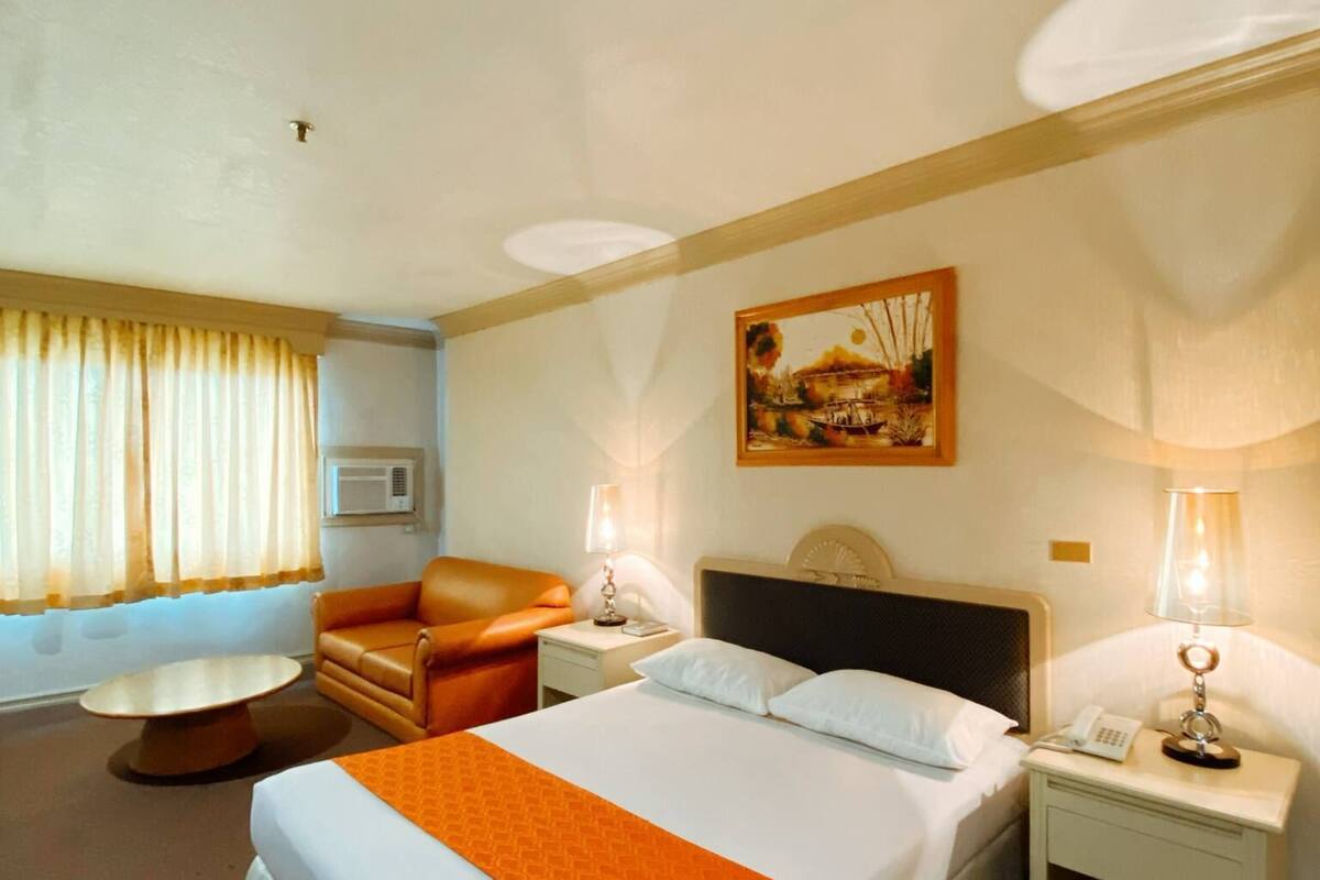 A hotel room with a large bed, a chair, a small table, and a painting on the wall.