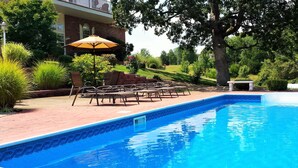 A swimming pool with lounge chairs and an umbrella, surrounded by greenery and a brick building. - Lafayette Grand Manor (Waynesville)