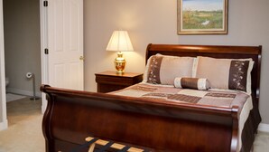 A bedroom with a wooden bed, a nightstand with a lamp, a framed landscape painting on the wall, and a door leading to another room. - Lafayette Grand Manor (Waynesville)