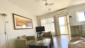 A living room with a sofa, a coffee table, a TV, and a ceiling fan. - Atlantic West Beach Apartments (West Beach)