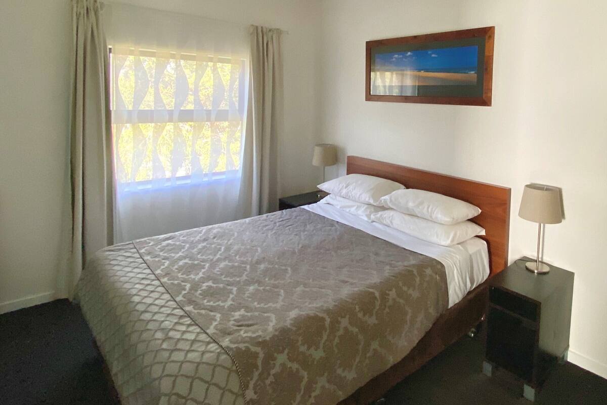 Ein Hotelzimmer mit einem Bett, einem Deckenventilator, einem Fenster mit Vorhängen, einem Nachttisch mit Lampe und einem Bild an der Wand.