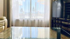 A living room with a glass coffee table, a sofa, a TV on a stand, and a large window with sheer curtains. - Atlantic West Beach Apartments (West Beach)