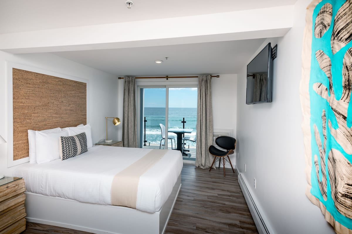 A hotel room with a large bed, a wooden headboard, a desk with a lamp, a TV mounted on the wall, and a view of the ocean through the sliding glass door.