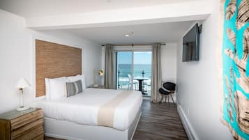 A hotel room with a large bed, a wooden headboard, a desk with a lamp, a TV mounted on the wall, and a view of the ocean through the sliding glass door.