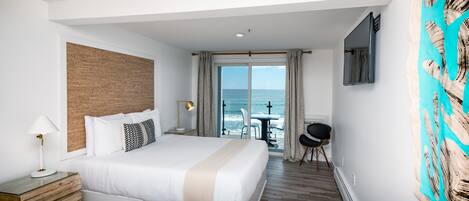 Habitación de hotel con una cama grande, cabecera de madera, escritorio con lámpara, televisor montado en la pared y vista al océano a través de la puerta corrediza de vidrio.