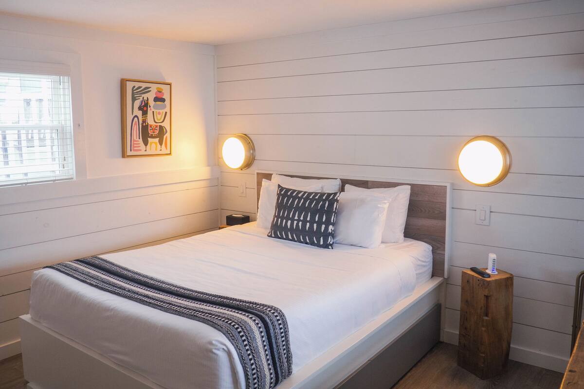 A neatly made bed with a patterned throw, two bedside lamps, a small wooden nightstand, and a framed picture on the wall.