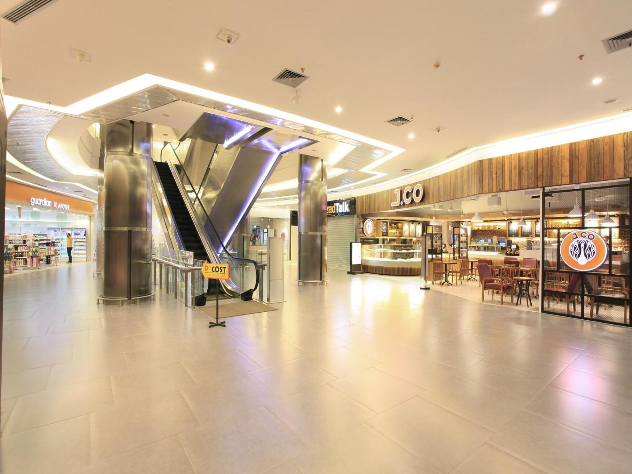 A spacious indoor area with a modern design, featuring a prominent escalator, a coffee shop named 'J.CO', and a store named 'Guardian'.