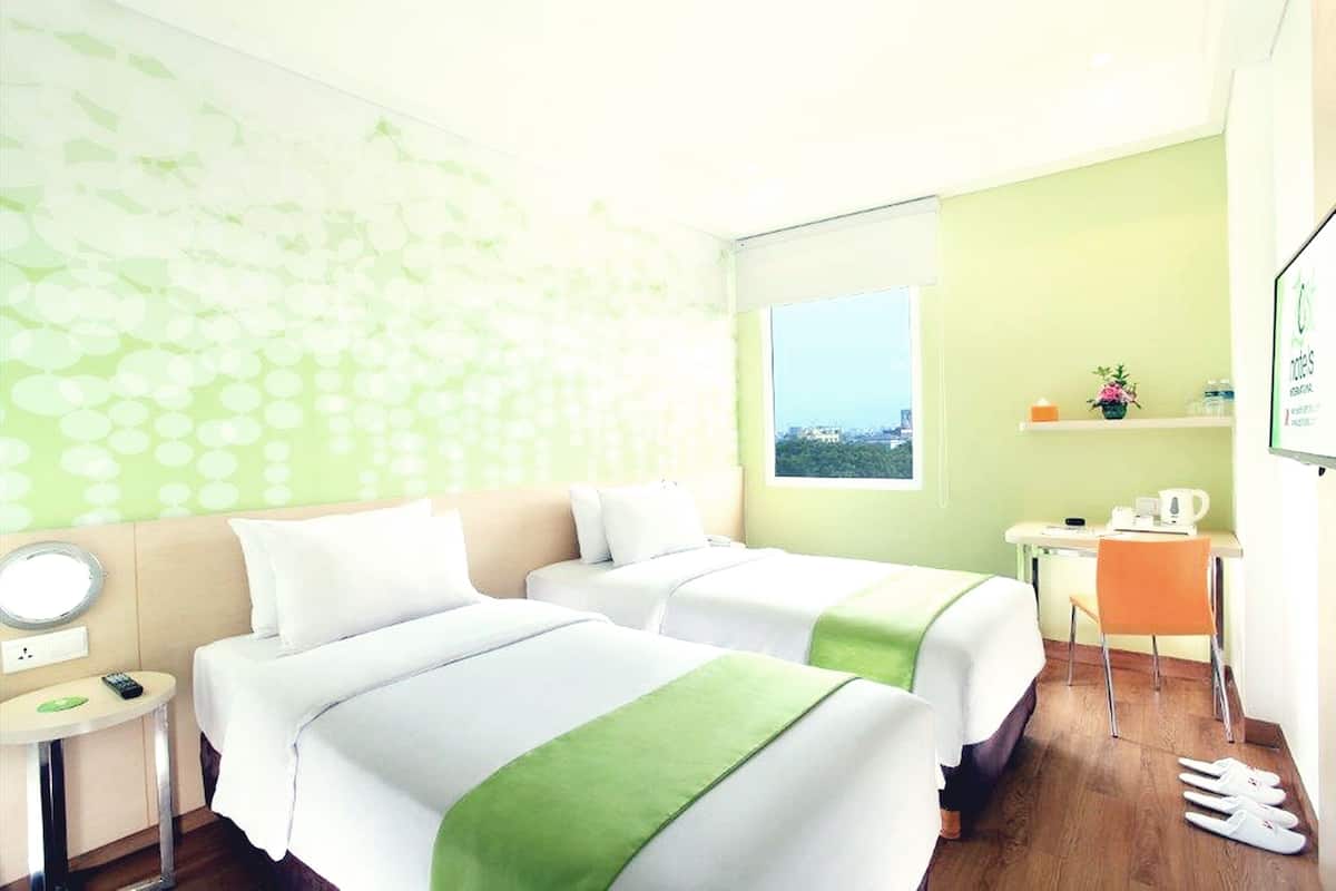 A hotel room with two beds, a desk, a chair, a small table, a mirror, and a window with a view.