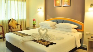 A hotel room with a neatly made bed, a decorative heart-shaped towel, a small table with a vase of flowers, and a chair with a cushion. - Ton Koon Hotel (Udon Thani)
