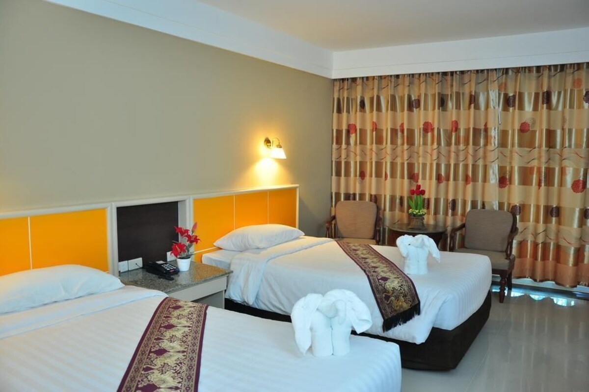 A hotel room with two beds, a bedside table with a lamp, a chair, and a small table with a vase.