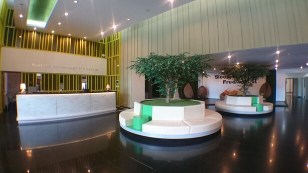 A modern lobby with a reception desk, green planters with trees, and a seating area. - Ton Koon Hotel (Udon Thani)