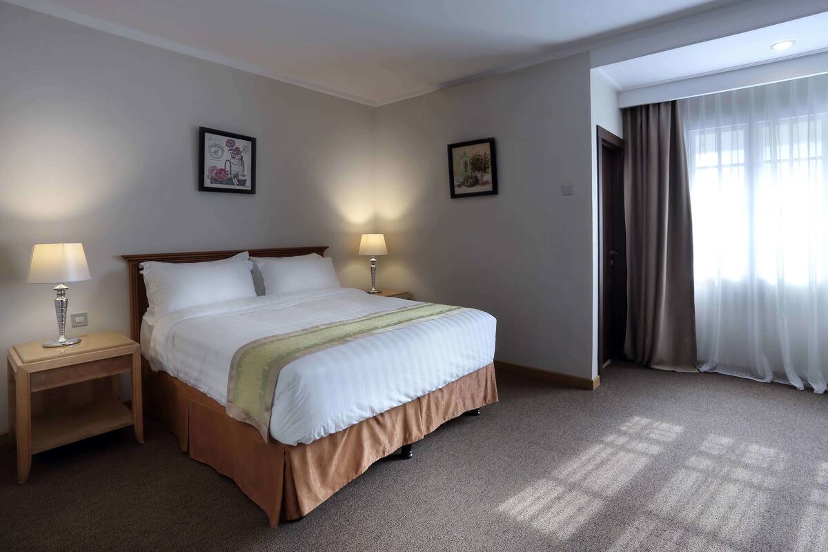 A hotel room with a bed, two bedside lamps, a nightstand, a window with curtains, and two wall-mounted pictures.