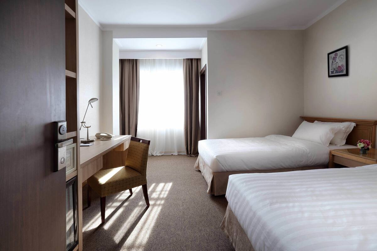 A hotel room with two beds, a desk with a chair, a lamp, and a window with curtains.
