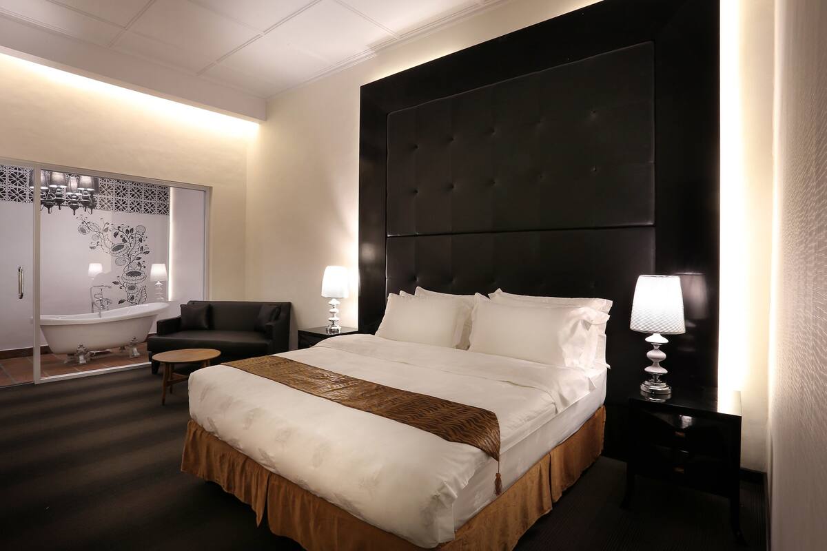A hotel room with a large bed, a black headboard, two bedside tables with lamps, a black sofa, a small round table, and a decorative mirror.