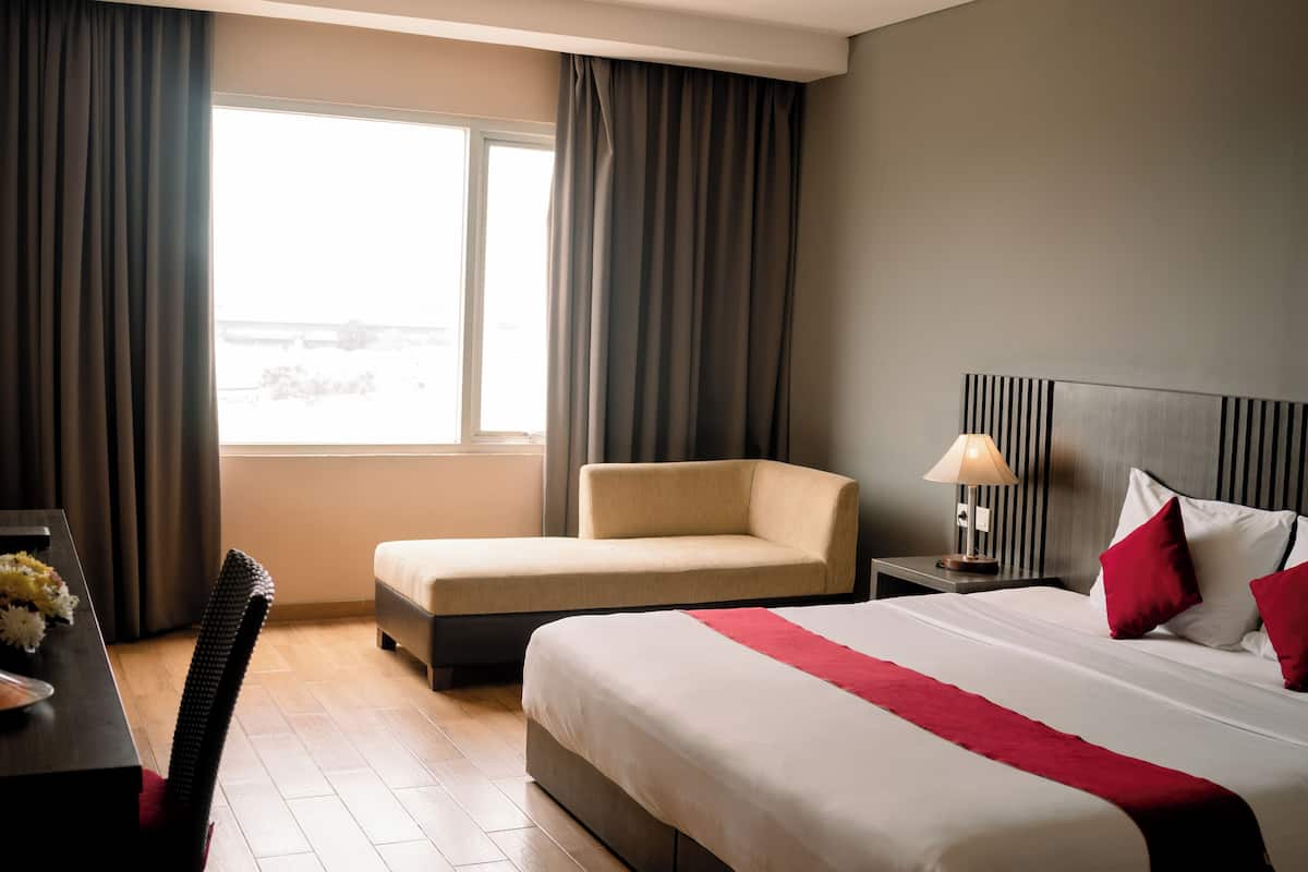 A hotel room with a bed, a sofa, a nightstand with a lamp, and a window with curtains.