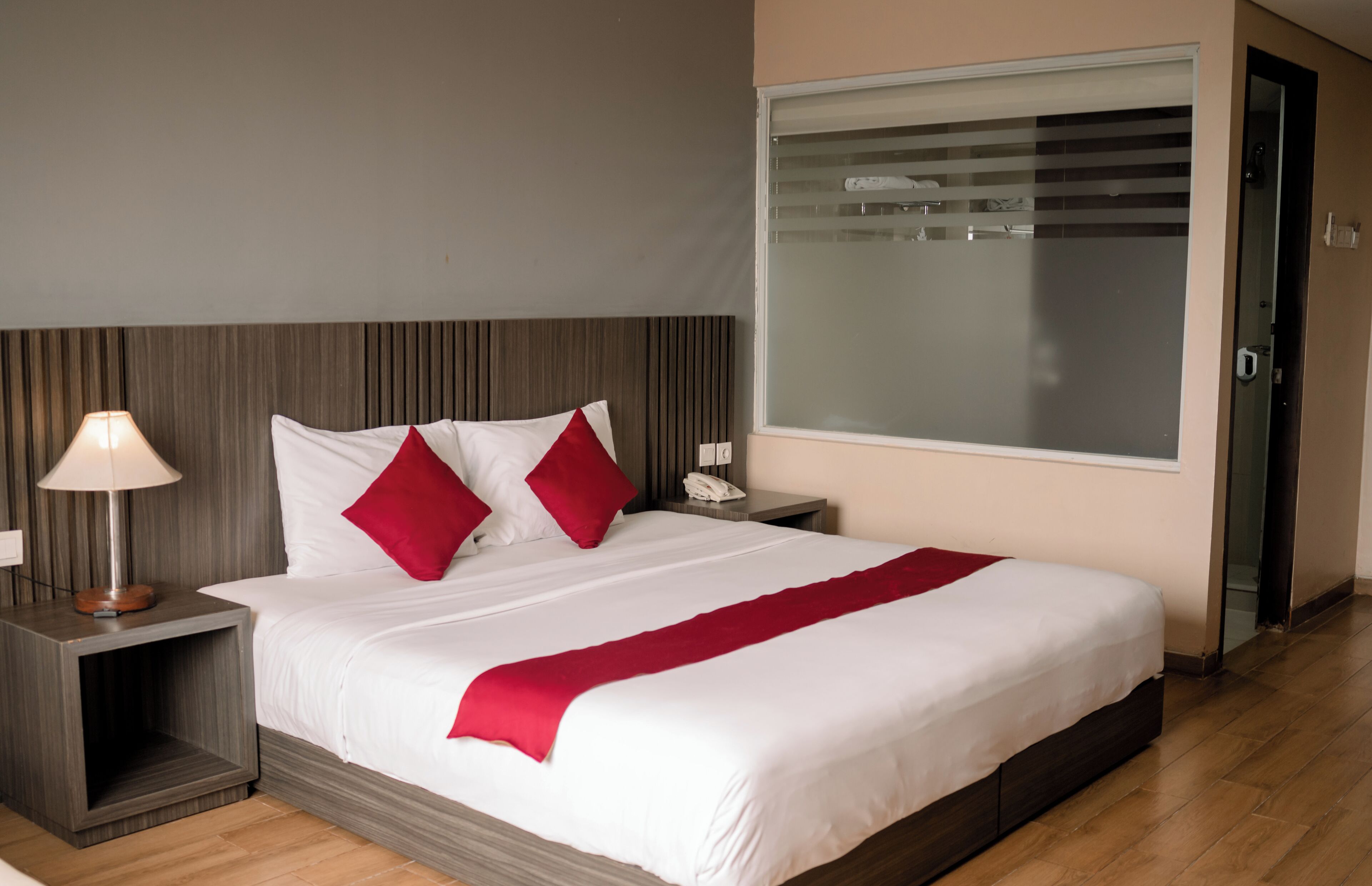 Una cama bien hecha con ropa de cama blanca y roja, una lámpara de noche y un teléfono sobre una mesita de noche de madera.
