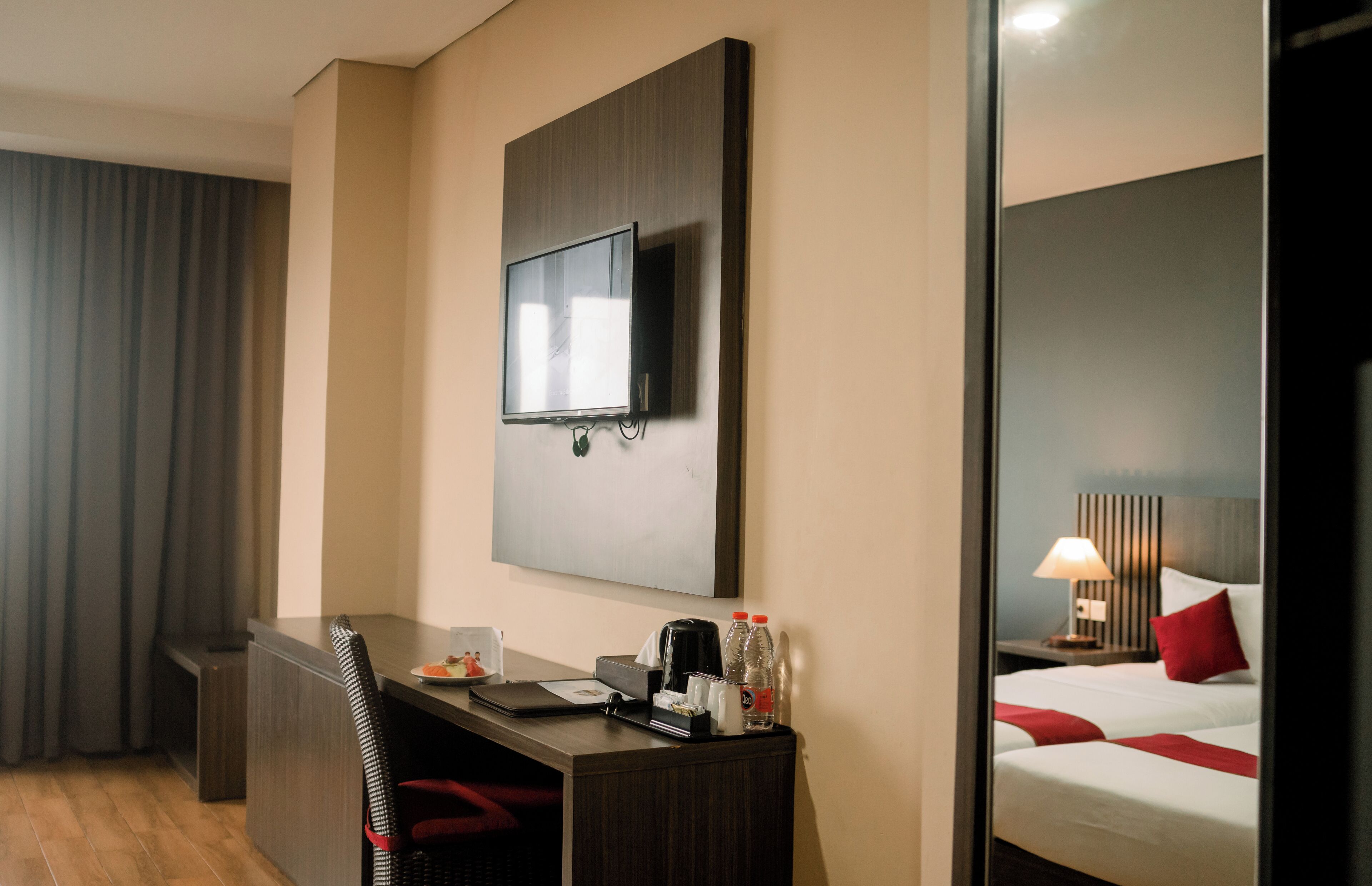 A hotel room with a desk, chair, bed, bedside table, lamp, and a TV mounted on the wall.