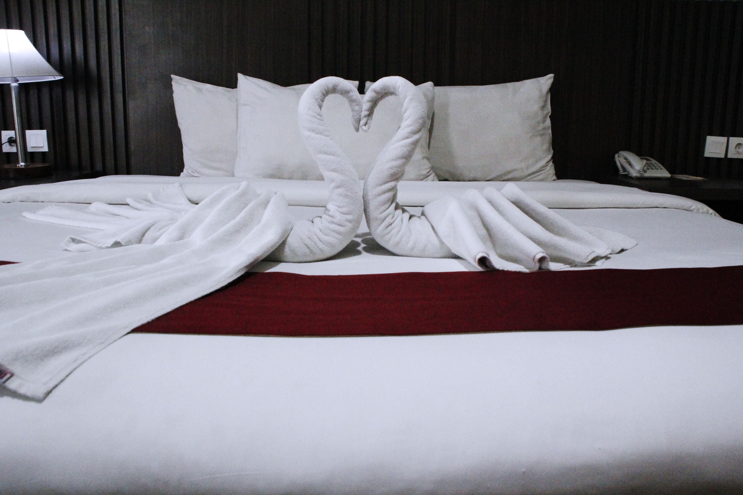 A neatly made bed with white linens and a red blanket. There is a heart-shaped towel arrangement on the bed, and a lamp is visible on the left side.