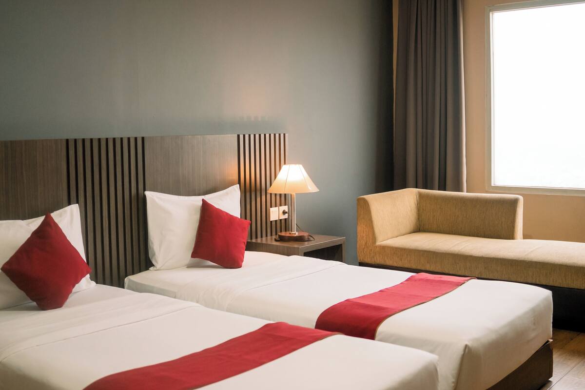 A hotel room with two beds, a nightstand with a lamp, a chair, and a large window.