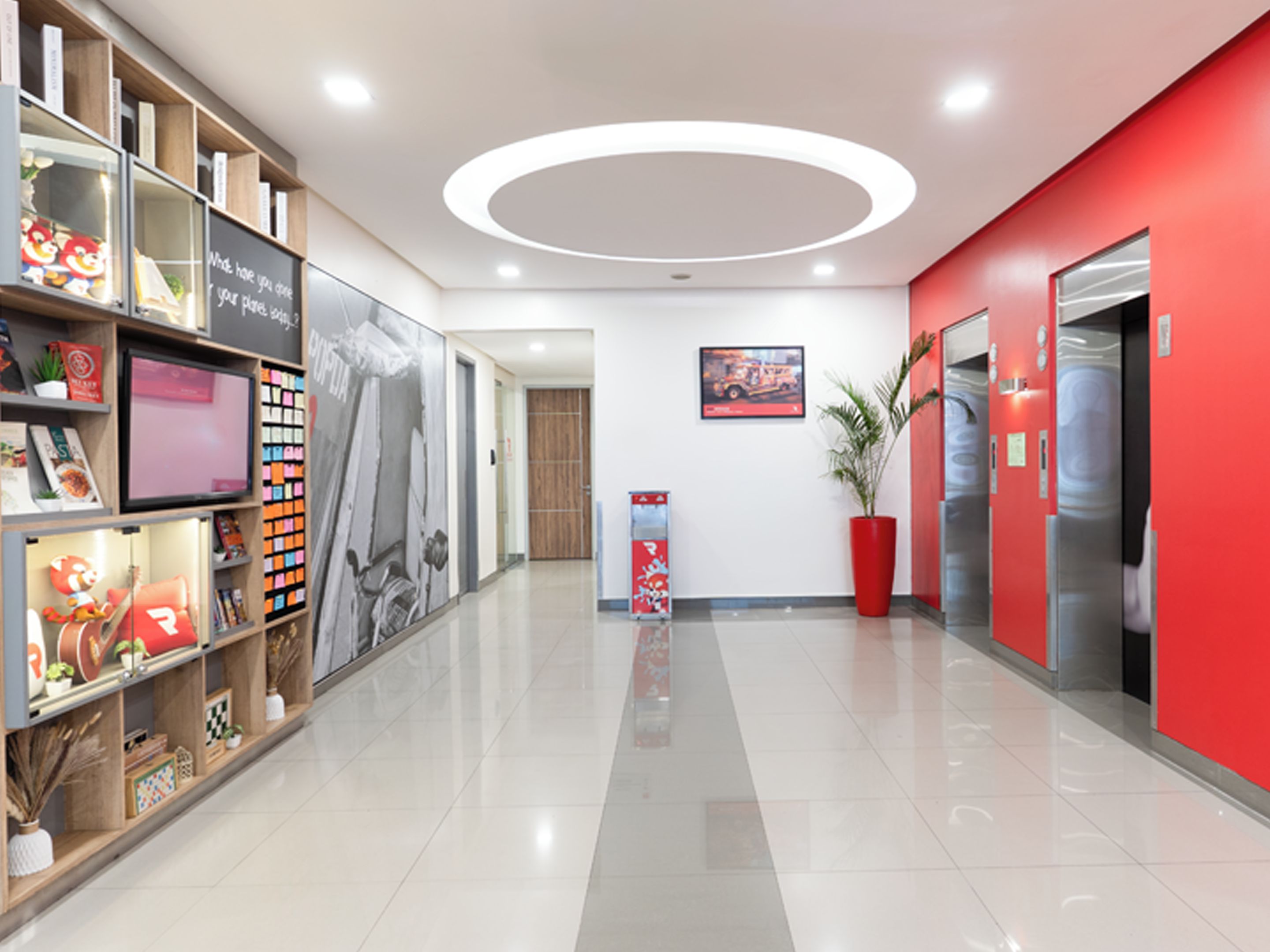 A modern hallway with a red accent wall, a large potted plant, and a digital display.