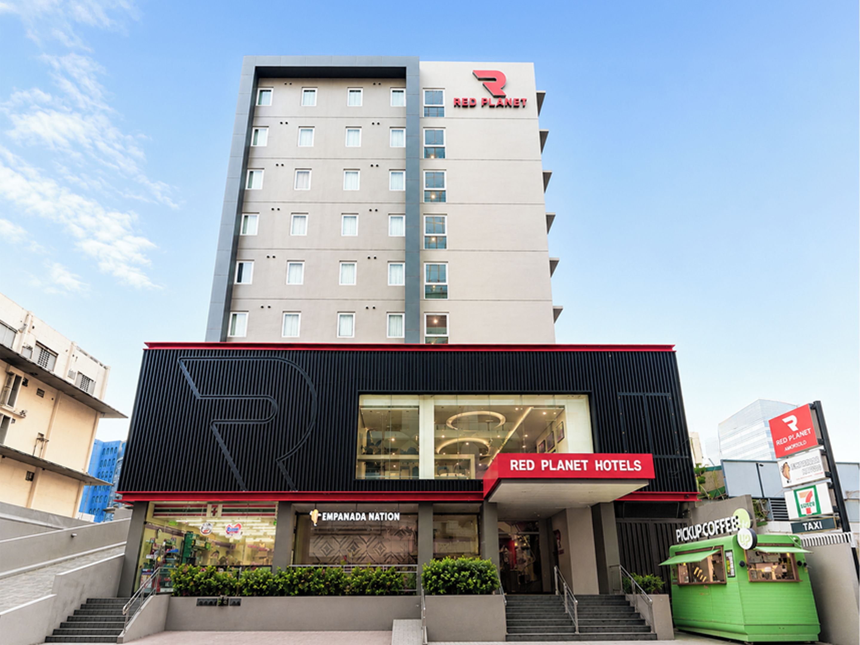 Un bâtiment moderne, sur lequel on peut voir une enseigne bien visible de la chaîne Red Planet Hotels, ainsi qu’un magasin 7-Eleven et un café au décor verdoyant.