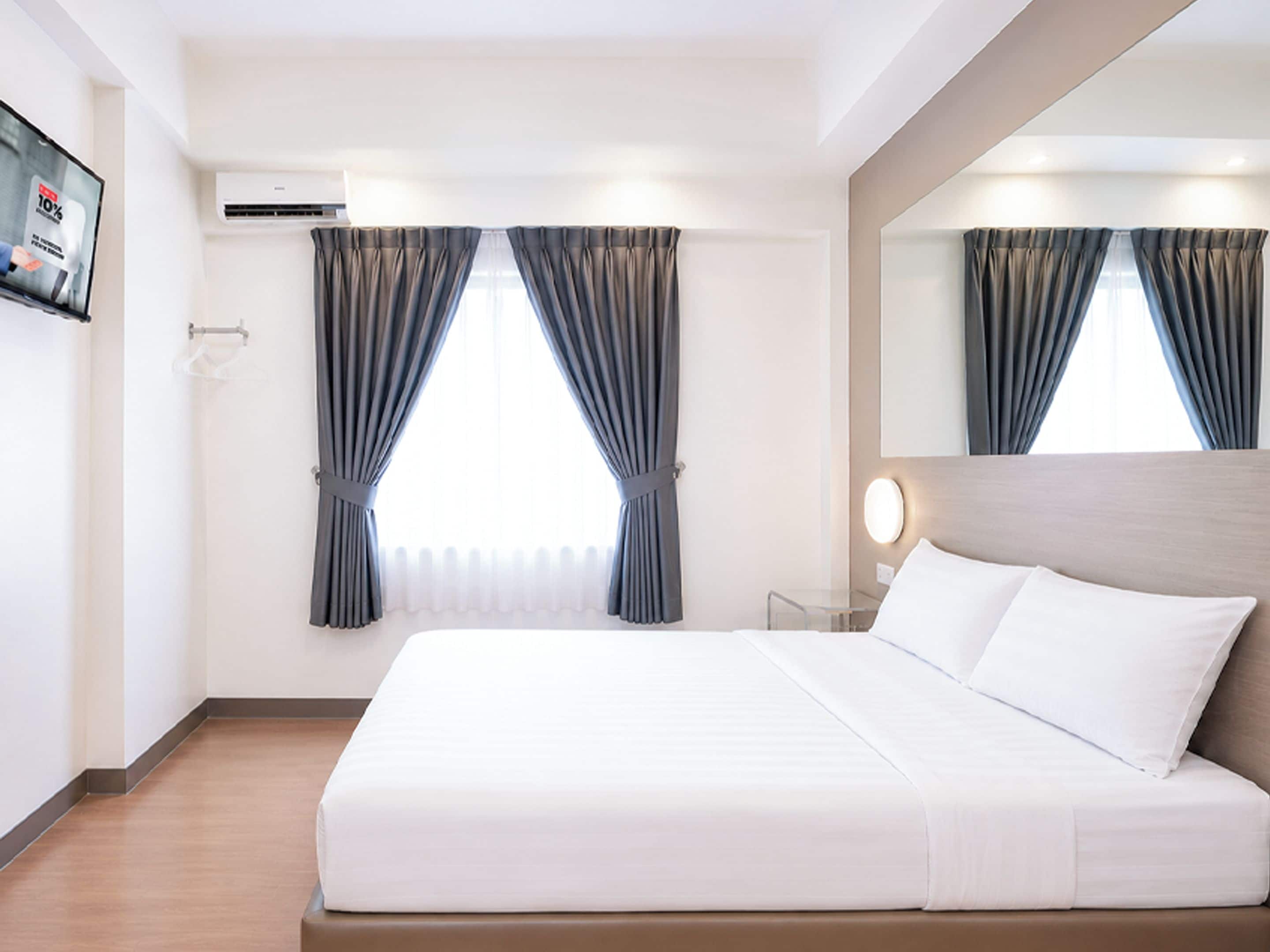 A hotel room with a bed, a TV, an air conditioning unit, and curtains.