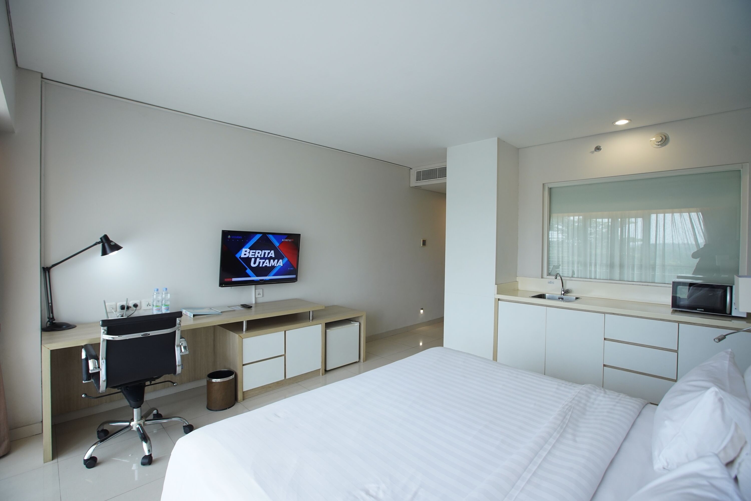 Una habitación de hotel moderna con una cama grande, un escritorio con computadora y un televisor de pantalla plana, una kitchenette pequeña y un espejo grande.