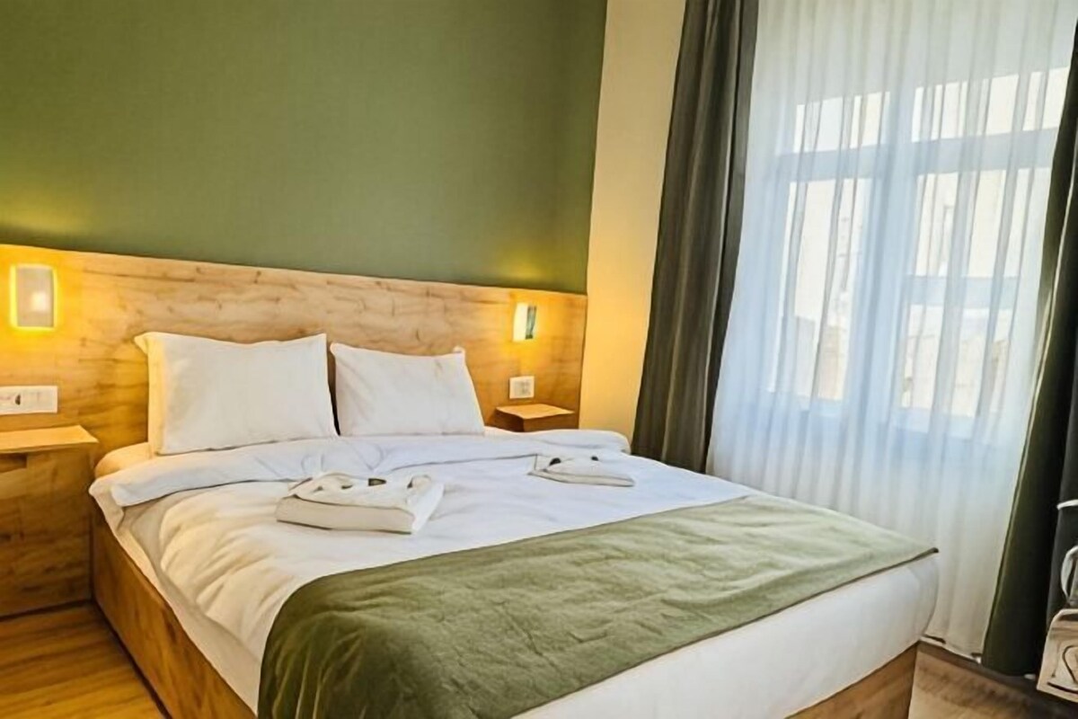 A hotel room with a wooden headboard, two bedside tables, a bed with white linens, and a green throw blanket.