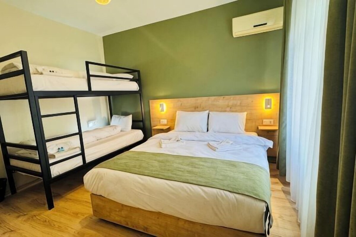 A room with bunk beds, a single bed, wooden flooring, a headboard with wall-mounted lights, and a window with curtains.