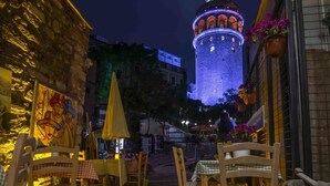 Uma área de estar ao ar livre com cadeiras e mesas de madeira, uma parede de pedra e um edifício ao fundo com luzes azuis e laranjas iluminadas.