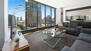 A modern living room with a large window, a glass coffee table, a grey sofa, and a kitchen area visible through the window. - Corporate Keys - Freshwater Place (Southbank)