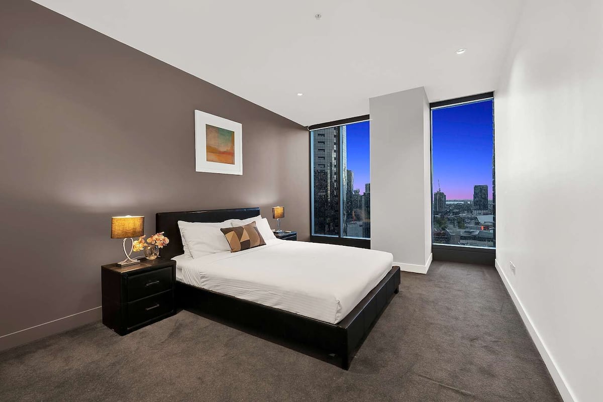 A modern bedroom with a large bed, nightstands, a city view, and a framed picture on the wall.