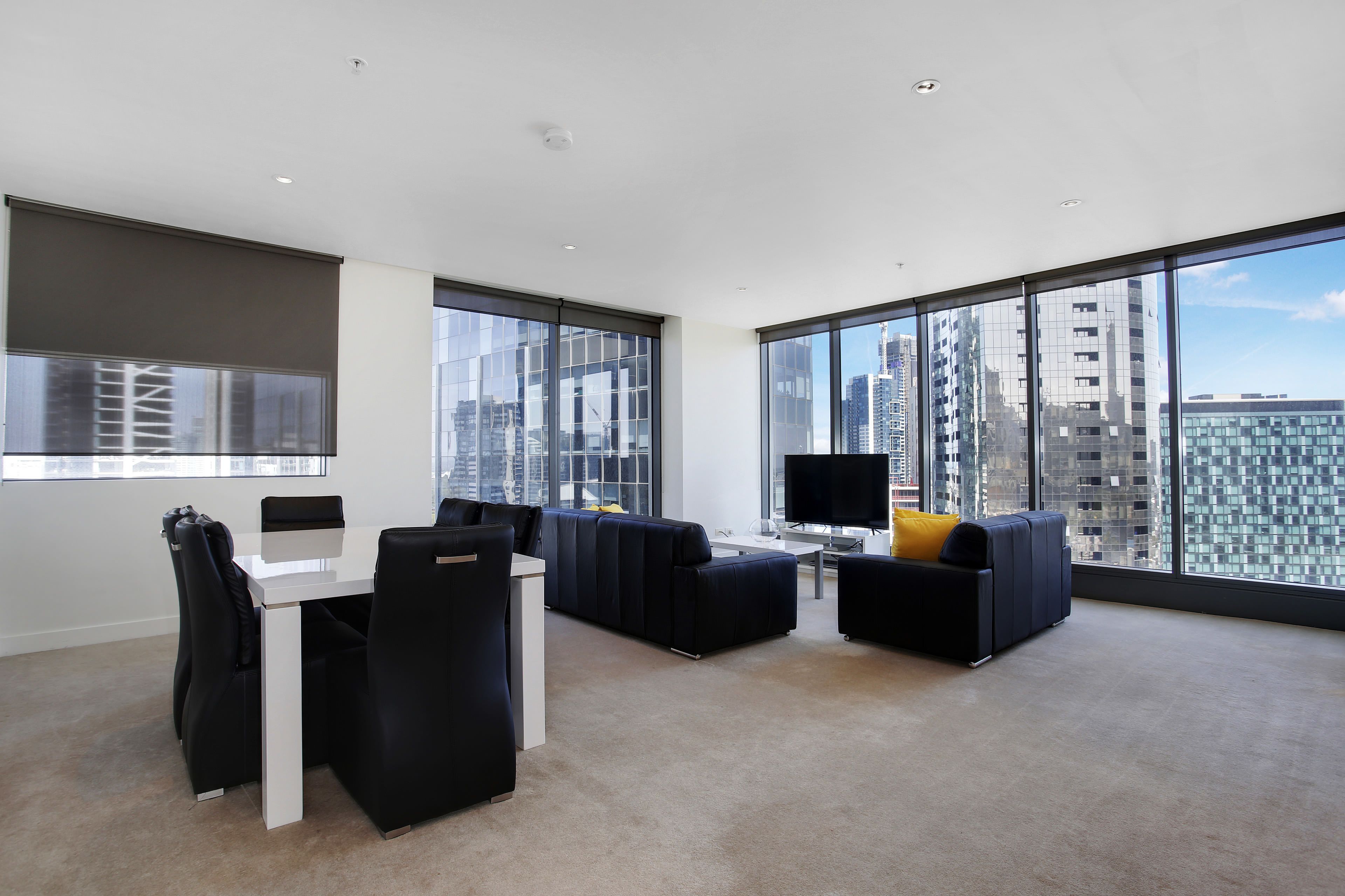 A modern living room with a white desk, black leather chairs, a black sofa with a yellow pillow, and large windows offering a city view.