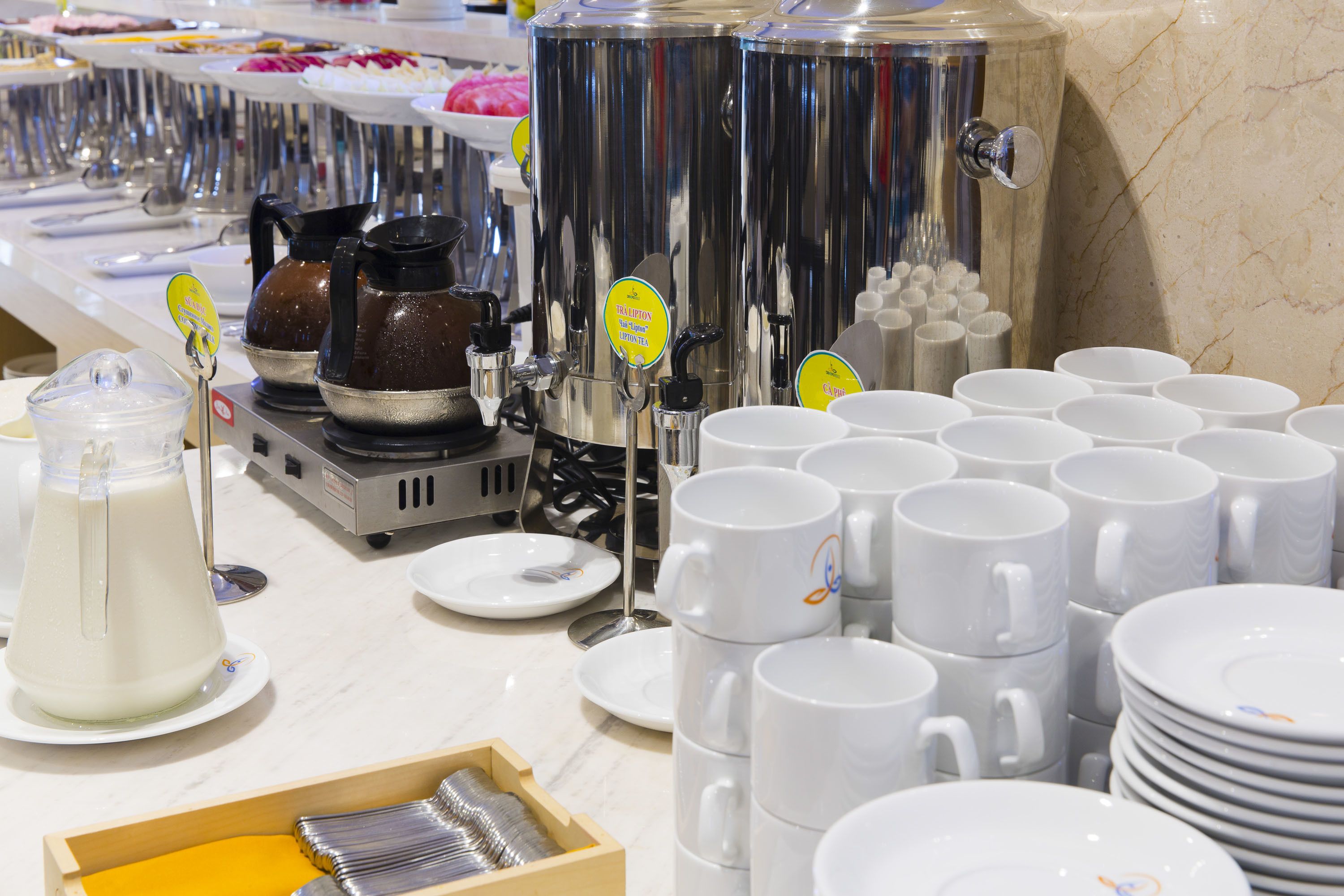 A buffet setup with multiple beverage dispensers, white mugs, and a milk dispenser.