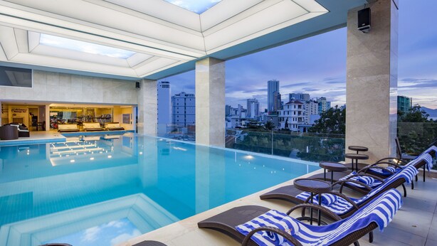 A modern indoor swimming pool with lounge chairs and a city view. - Dendro Gold Hotel (Nha Trang)