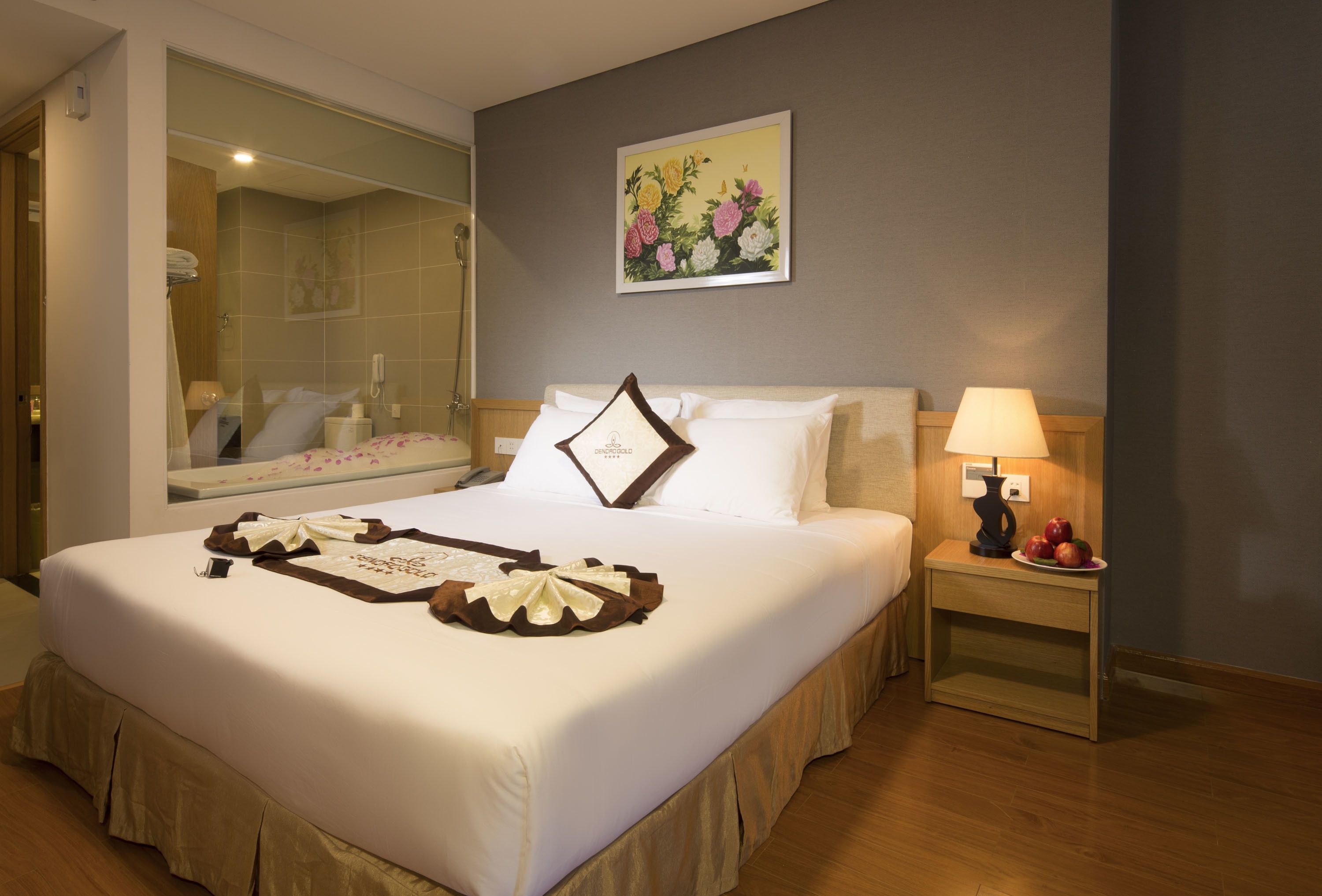 A neatly made bed with a decorative pillow, a bedside table with a lamp and fruit, and a large mirror reflecting the room.