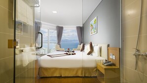 A hotel room with a large bed, a bedside table with a phone, a glass shower enclosure, and a view of the sea. - Dendro Gold Hotel (Nha Trang)