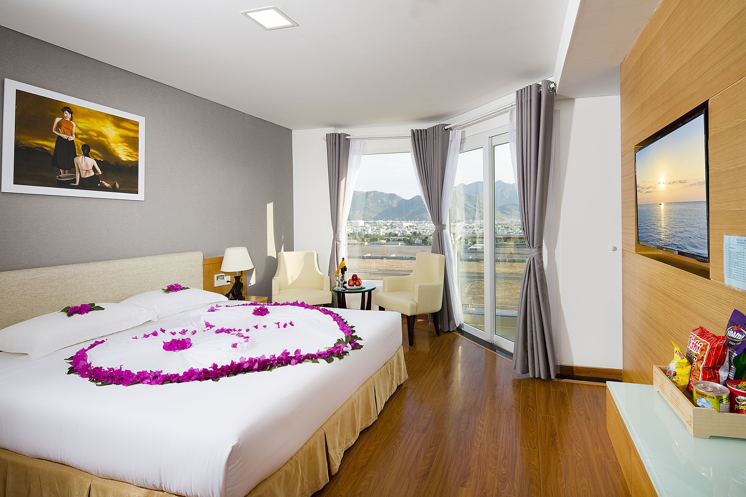 A hotel room with a large bed, a desk, a chair, a view of the city, and a painting on the wall.