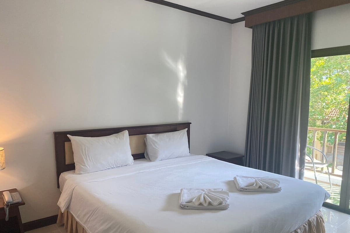 Quarto de hotel com uma cama grande, uma mesa de cabeceira com abajur e vista para o verde pela janela.
