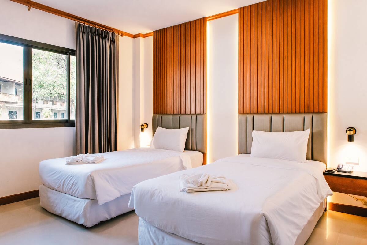 Quarto de hotel com duas camas, uma janela com cortinas, painéis de madeira nas paredes e uma mesa de cabeceira com abajur.