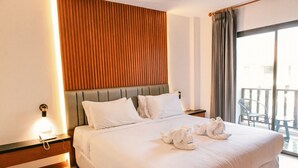 A hotel room with a large bed, a wooden accent wall, a nightstand with a lamp, and a view of the outside balcony. - President Hotel (Udon Thani)