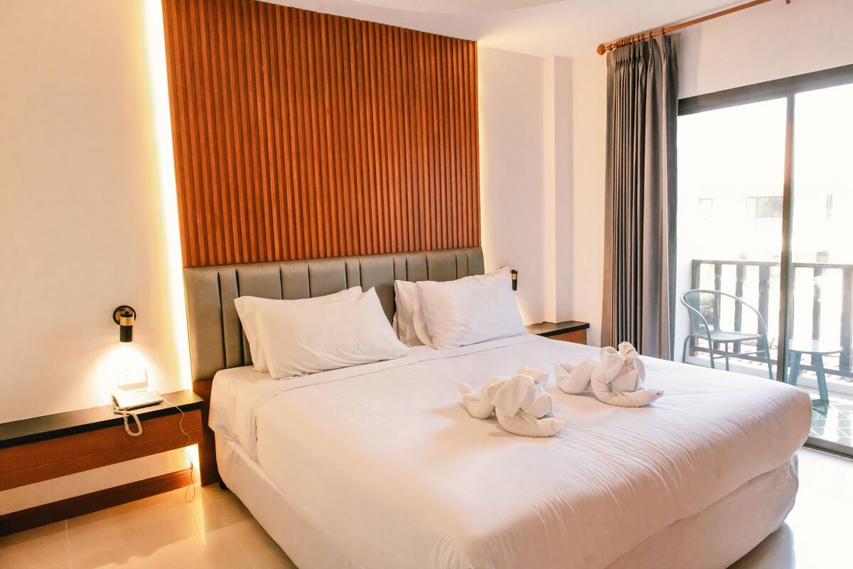 Quarto de hotel com uma cama grande, uma parede de destaque em madeira, uma mesa de cabeceira com abajur e vista para a varanda.