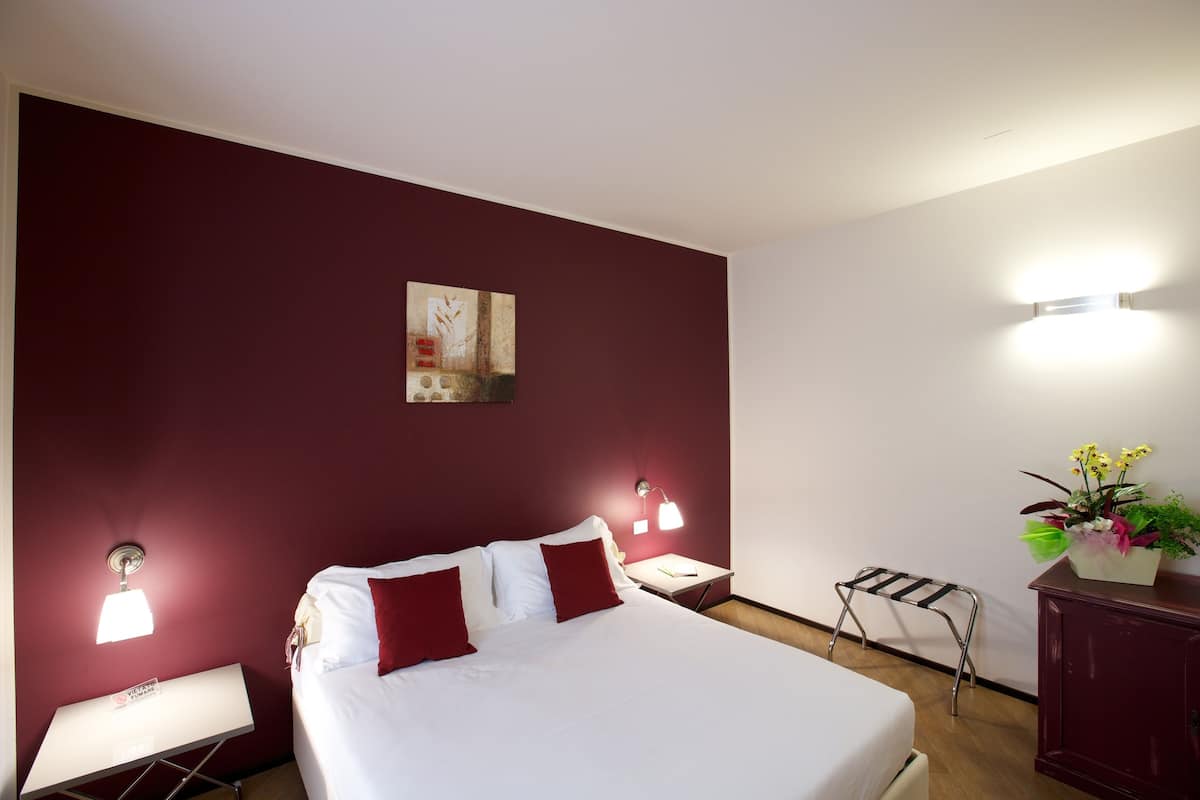 Une chambre d’hôtel avec un grand lit, deux tables de chevet, une petite table d’appoint, une lampe fixée au mur, un petit tabouret et une plante en pot.