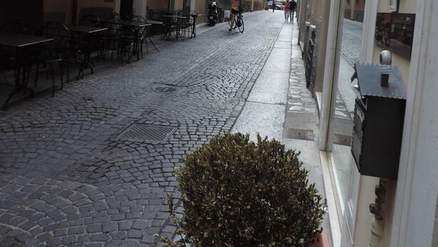 Una calle adoquinada flanqueada por edificios, con mesas al aire libre y una maceta en primer plano.